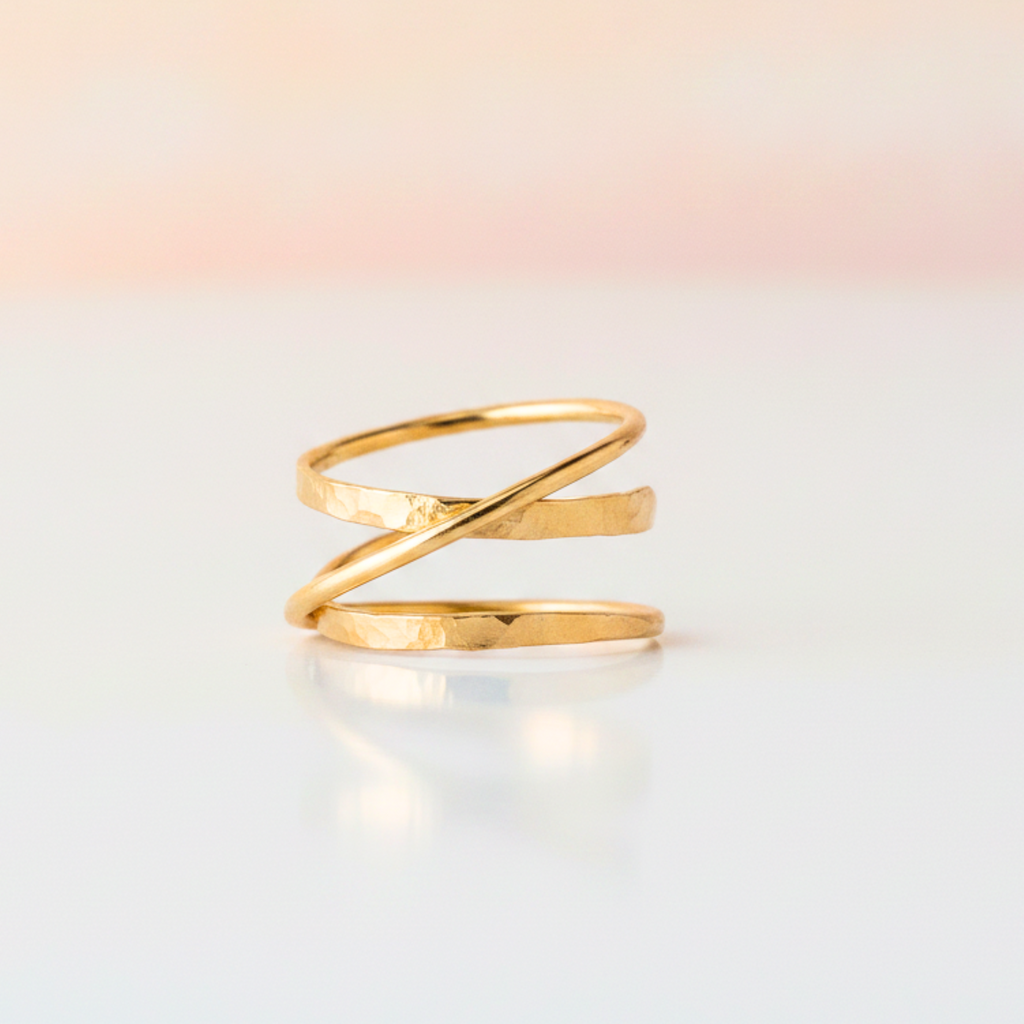 A gold-plated open wrap ring with a hammered texture, resting on a reflective white surface against a soft, warm gradient background.